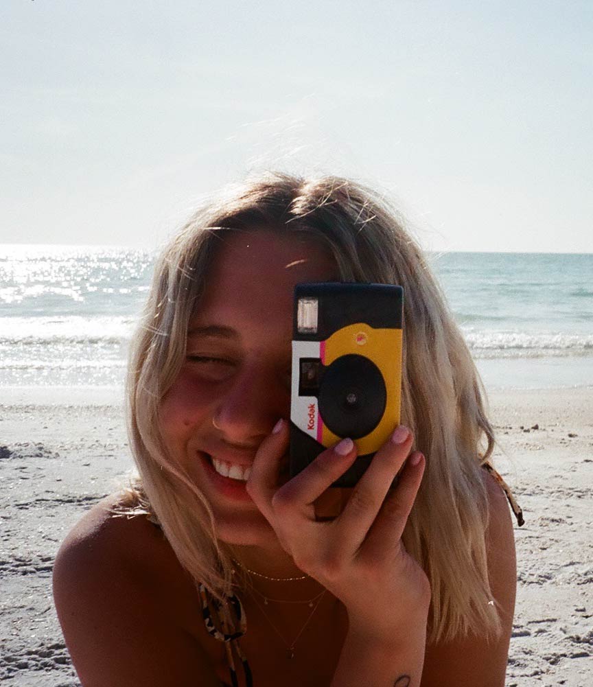 Aprille on a beach holding up a disposable camera, taking a photo.